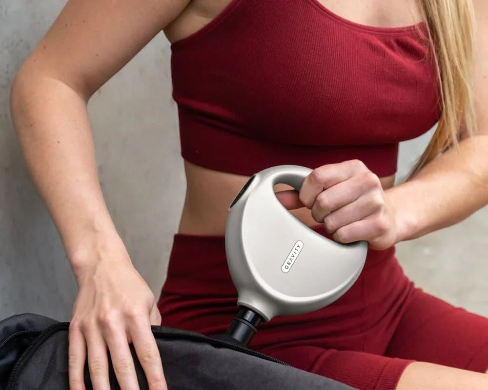 BirdieBox Gravity Move Heated Mini Massager in light grey, held by a woman in a maroon sports bra and shorts, next to a black gym bag.