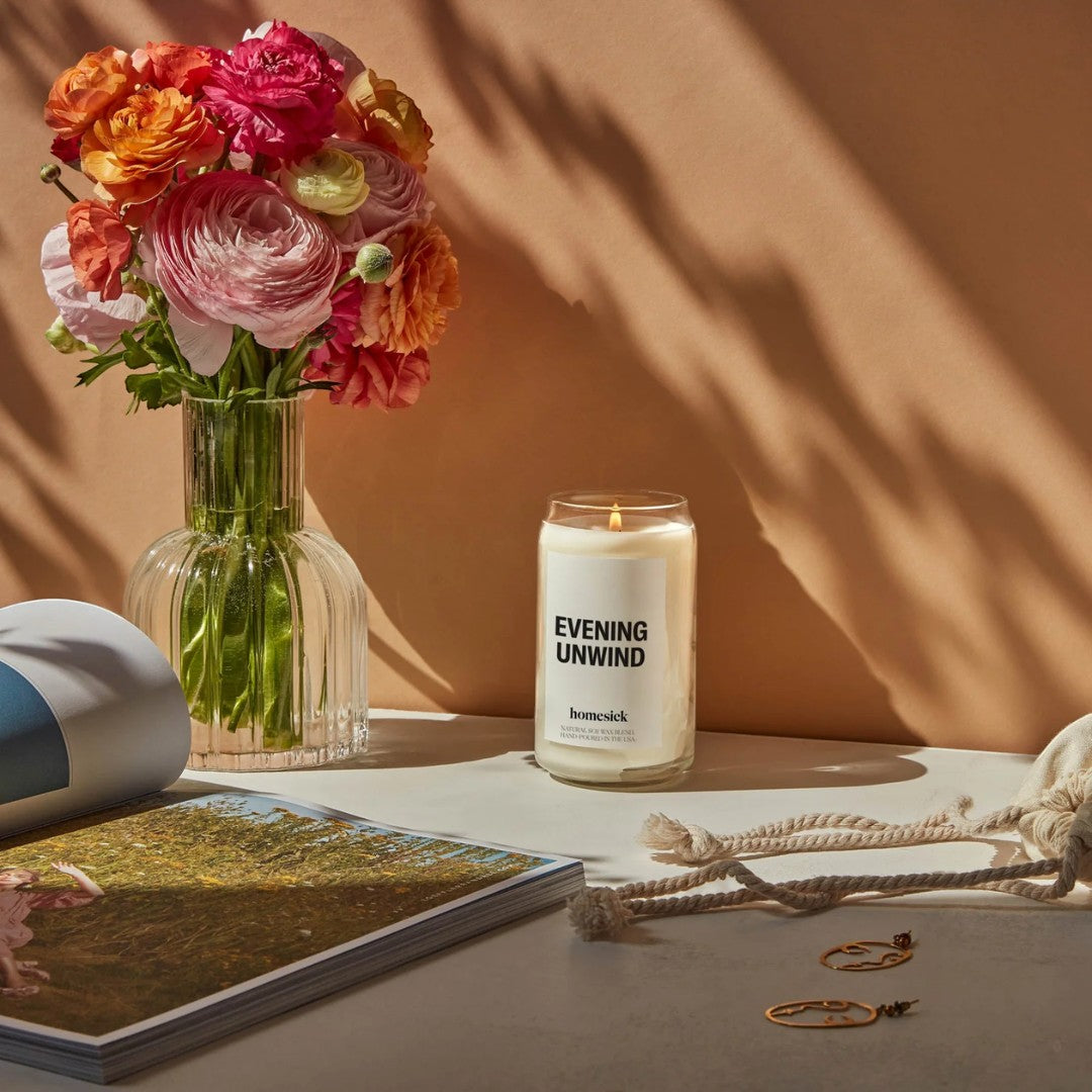 BirdieBox Homesick Evening Unwind candle in a glass jar with a white label, lit and placed next to a vase of flowers.