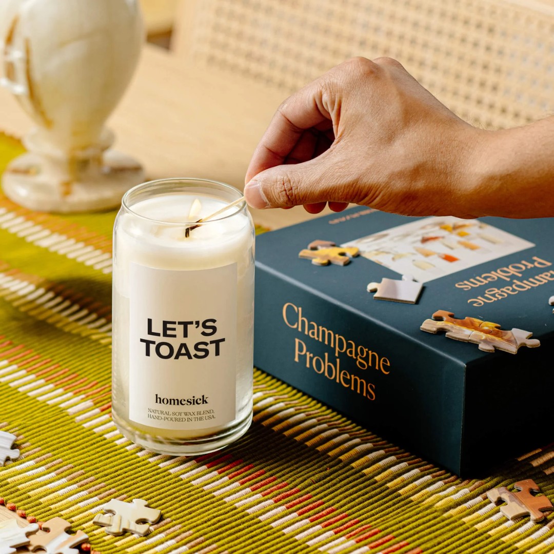 BirdieBox Homesick Let's Toast candle with champagne grape scent, lit by a hand holding a match. Next to a puzzle box.