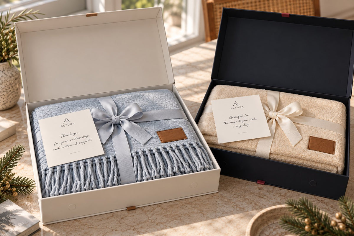 Two blanket gift sets: a fringed herringbone throw in a white magnetic box and a plush throw in a black box, each with a bow and note card (sample branding)