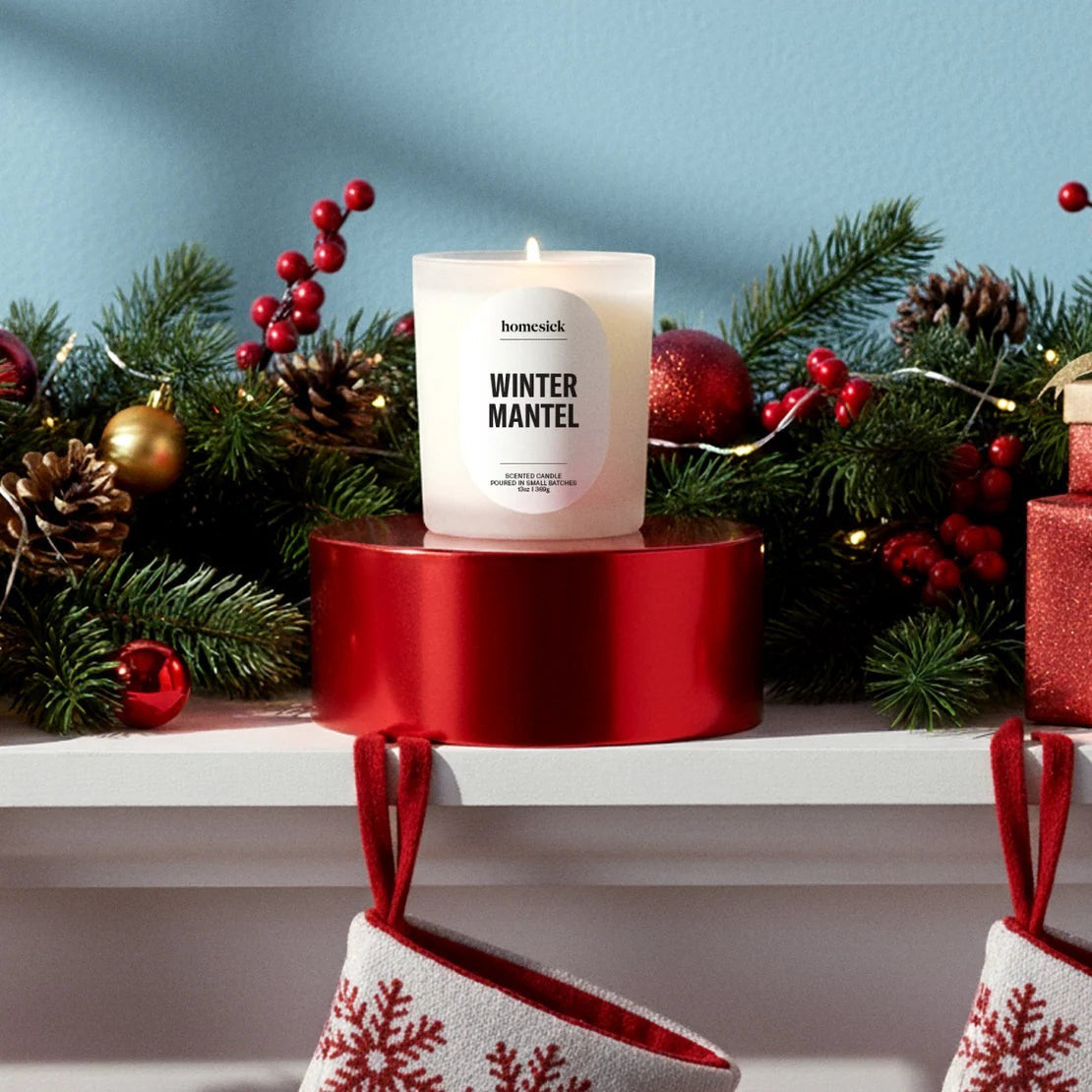 BirdieBox Homesick Winter Mantel scented candle in a frosted glass jar with a white label, surrounded by festive greenery, red ornaments, and Christmas stockings.